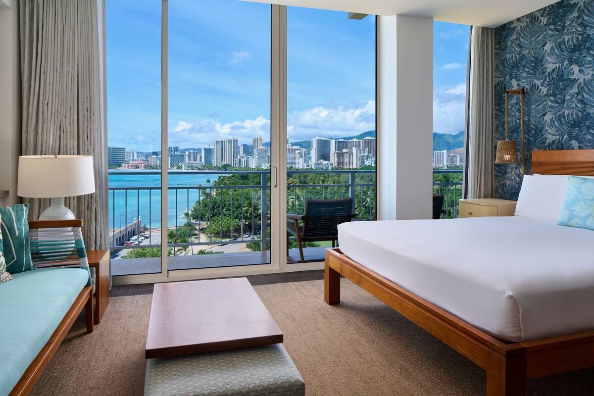 Habitación de hotel con una cama grande, un sofá, una mesita y vistas a la ciudad y la playa.