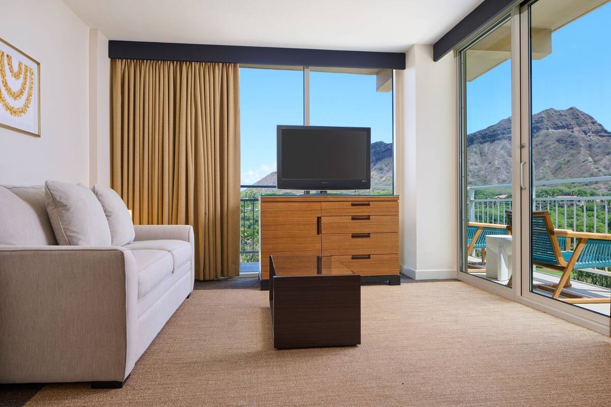 Una sala de estar moderna con un sofá, un televisor de pantalla plana sobre un mueble de madera y vistas a la montaña a través de amplios ventanales.