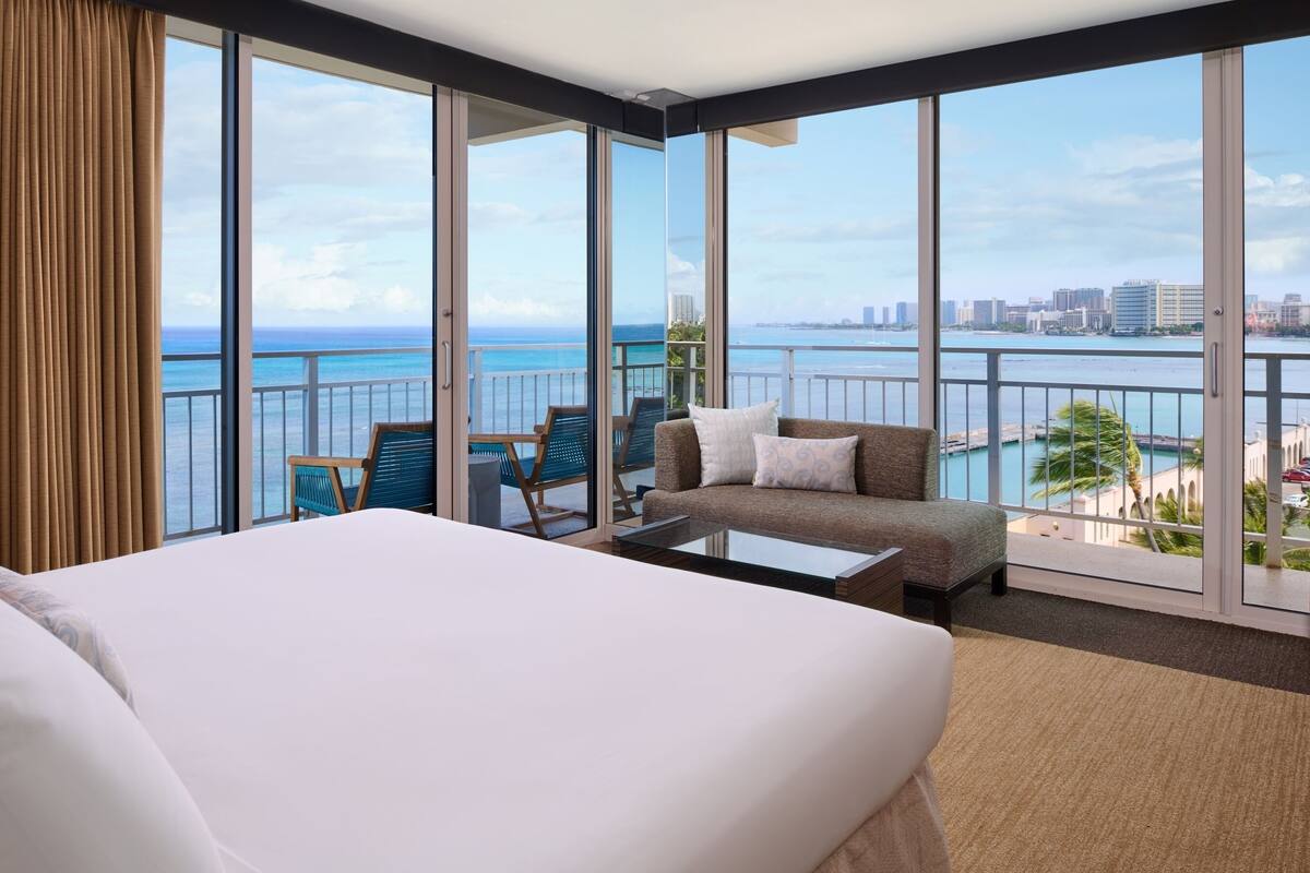 Habitación de hotel con una cama grande, un sofá, una mesa de centro y vistas al océano y al horizonte urbano a través de ventanales.