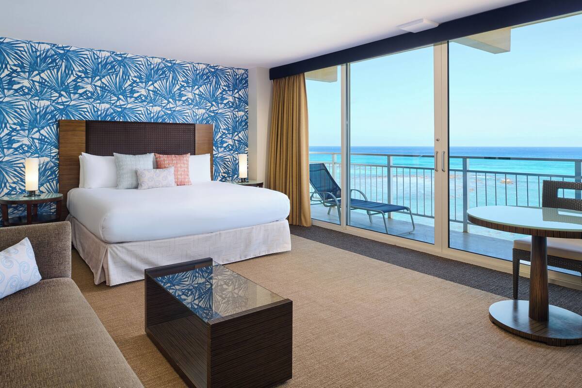 Habitación de hotel con una cama grande, un sofá, una mesita de centro de vidrio y un balcón con vista al océano.