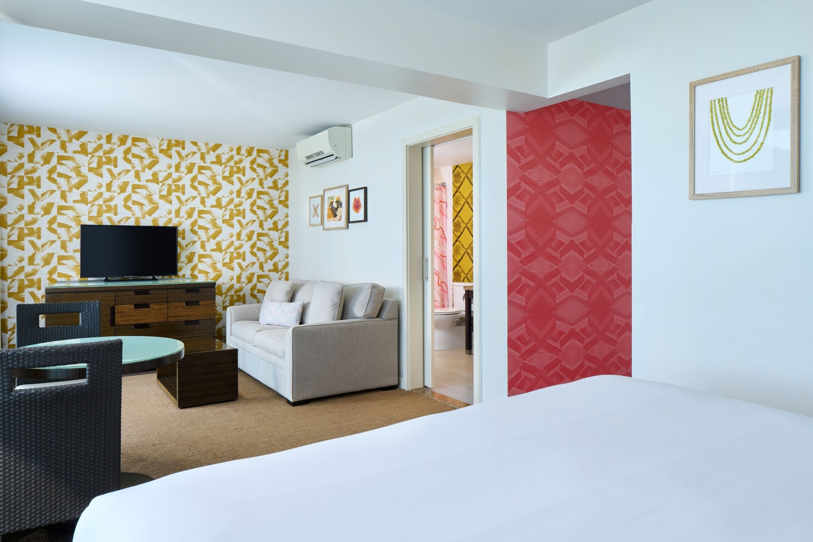A hotel room with a bed, a sofa, a TV, a dining table, and chairs. There is a red patterned wall and a yellow patterned wall.