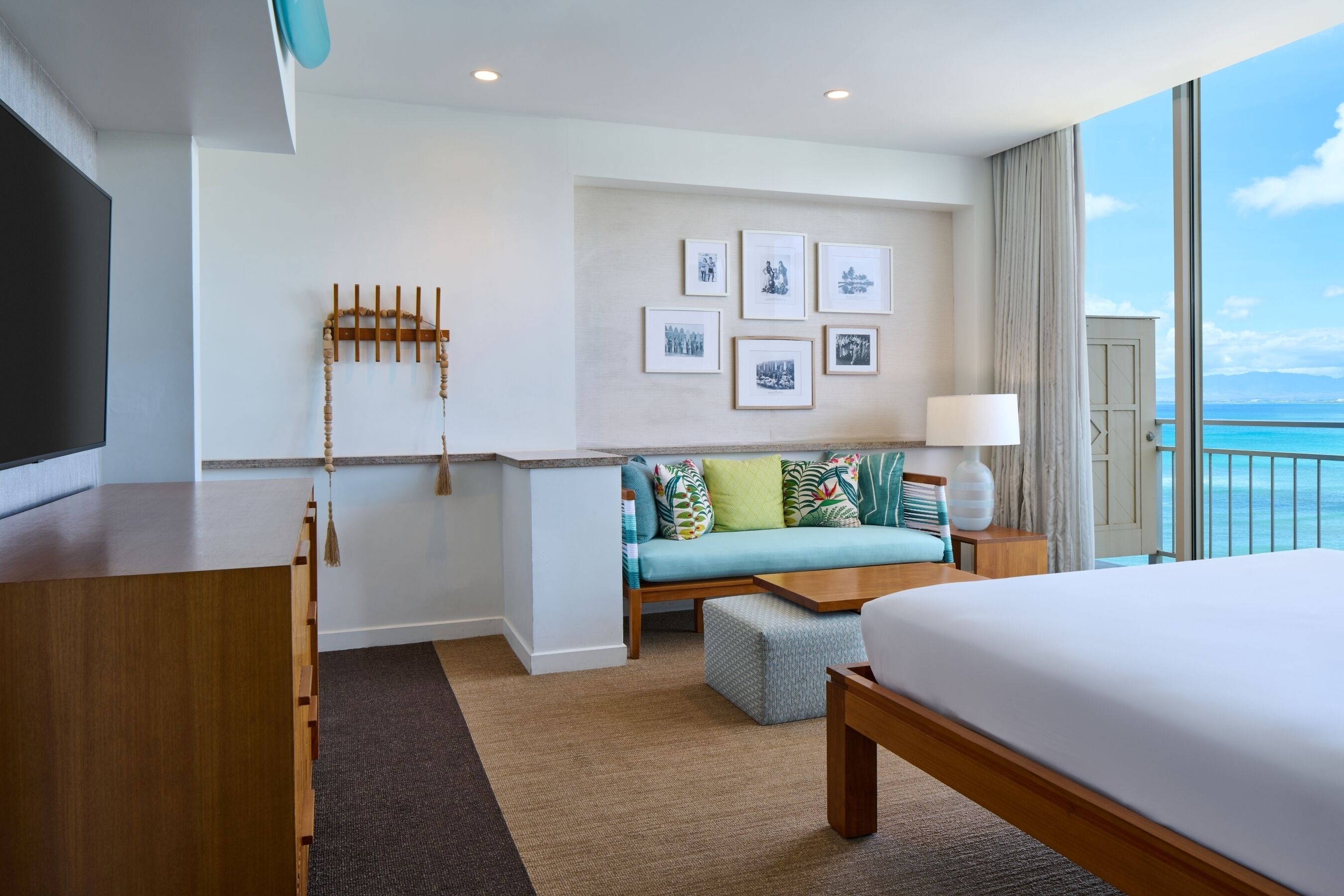 A hotel room with a bed, a sofa, a TV, a desk, and a balcony with a view of the ocean.