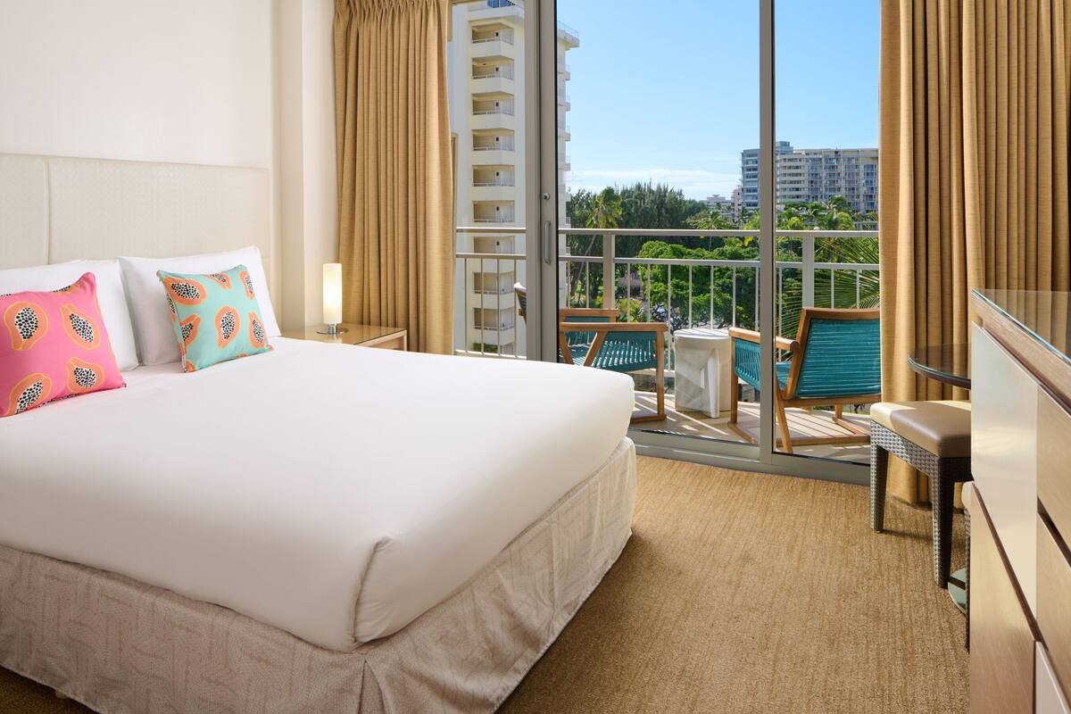 Habitación de hotel con una cama grande, un balcón con vistas a palmeras y edificios, y una zona de estar con una silla y una mesa.