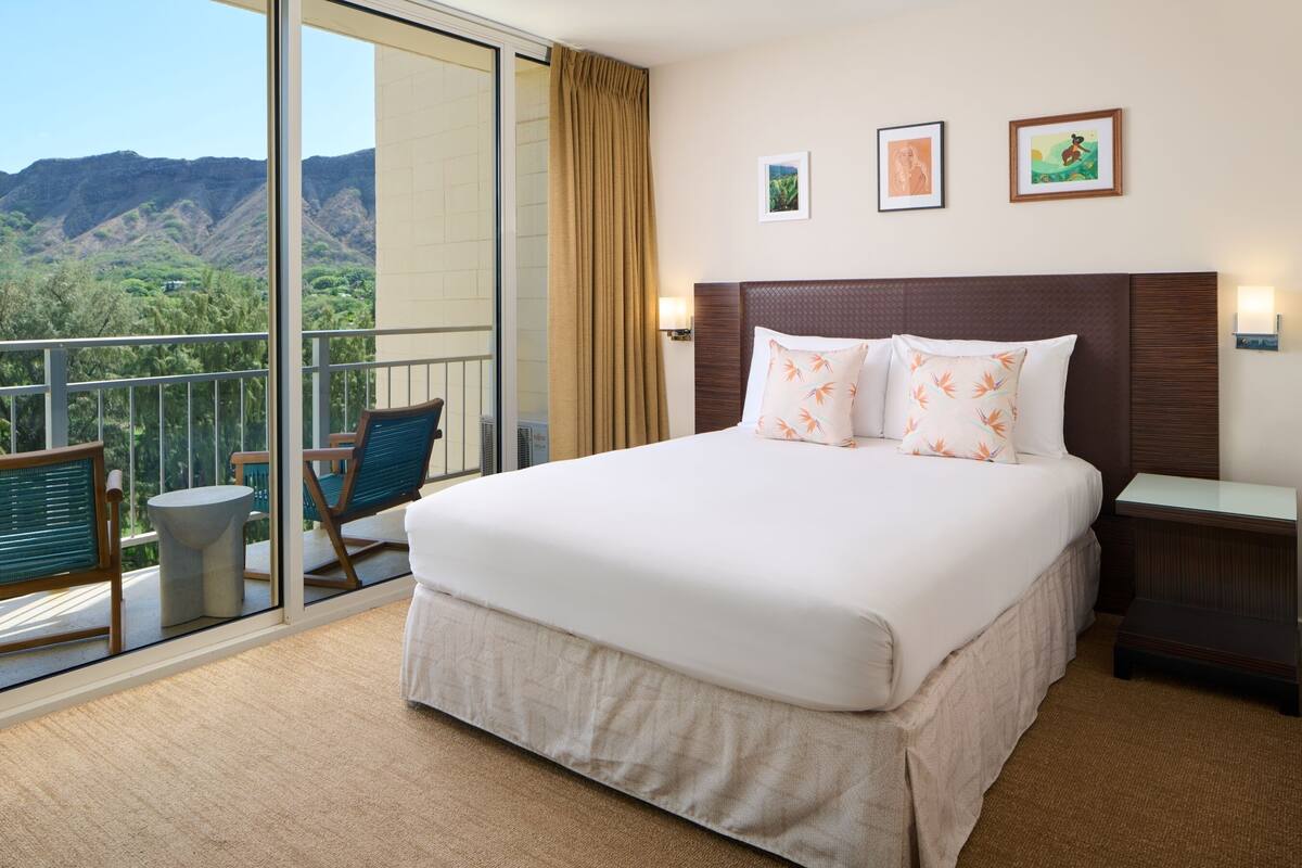 Habitación de hotel con una cama grande, un balcón con vistas a montañas y una pequeña mesa con sillas.