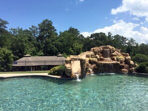 A clear pool with a rock waterfall feature, surrounded by lush greenery and a covered pavilion.