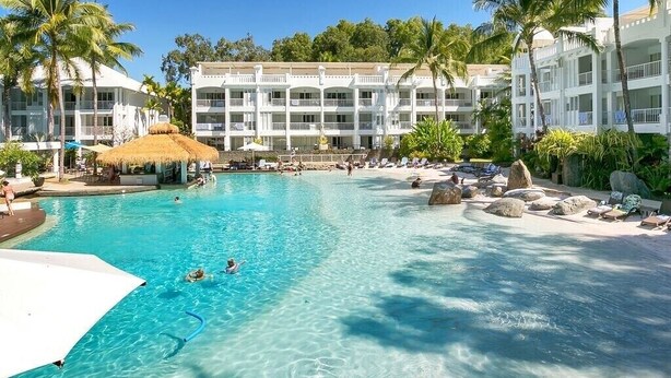 Uma piscina de resort com água azul cristalina, cercada por palmeiras e um edifício com várias varandas.