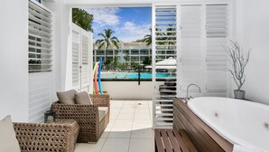 Uma casa de banho moderna com uma banheira grande, móveis de vime e vista para uma piscina e palmeiras.