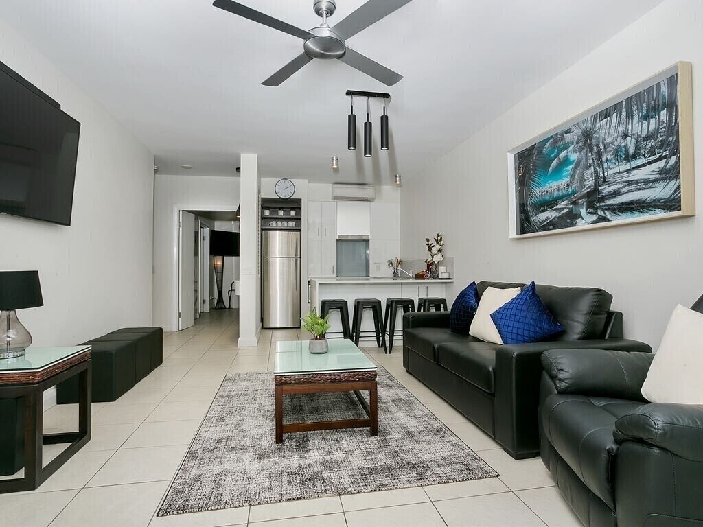 A modern living room with a black leather sofa, a coffee table, and a TV. There is a kitchen area in the background with a refrigerator and a dining table.