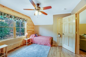 A cozy bedroom with a wooden ceiling fan, a bed with a patterned blanket, a nightstand with a lamp, and a view of trees through large windows. - Gorgeous Lakefront Home with a Fire Pit, Two Living Areas, and a Sunroom (Chestertown)