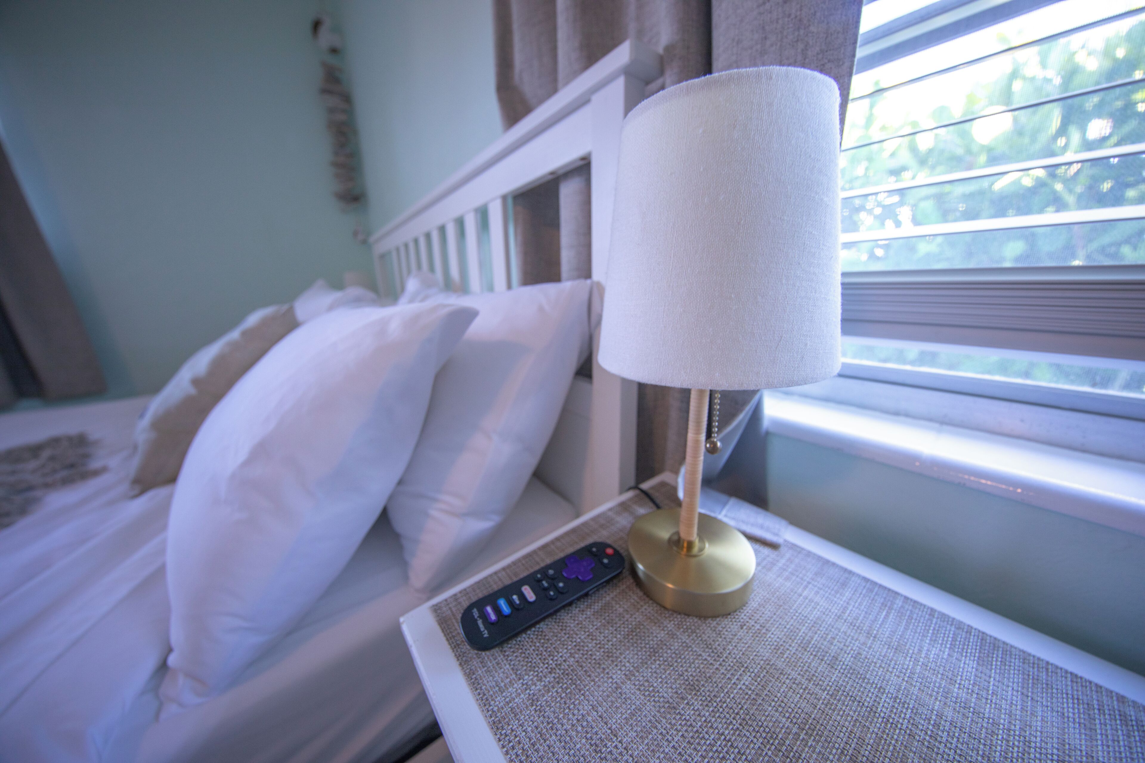 A neatly made bed with white linens and pillows, a bedside table with a lamp and remote control, and a window with blinds.