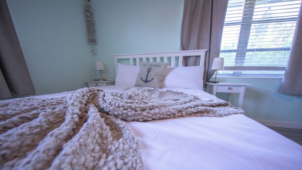 A neatly made bed with a knitted blanket, a pillow with an anchor design, and a nightstand with a lamp. - Centrally Located Charming Beach Suite in Sarasota (Sarasota)