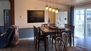 A dining area with a wooden table, chairs, a chalkboard with a welcome message, pendant lights, and a view of the outdoors. - Enjoy Lake Life, A Great Minnesota Getaway! (Henning)