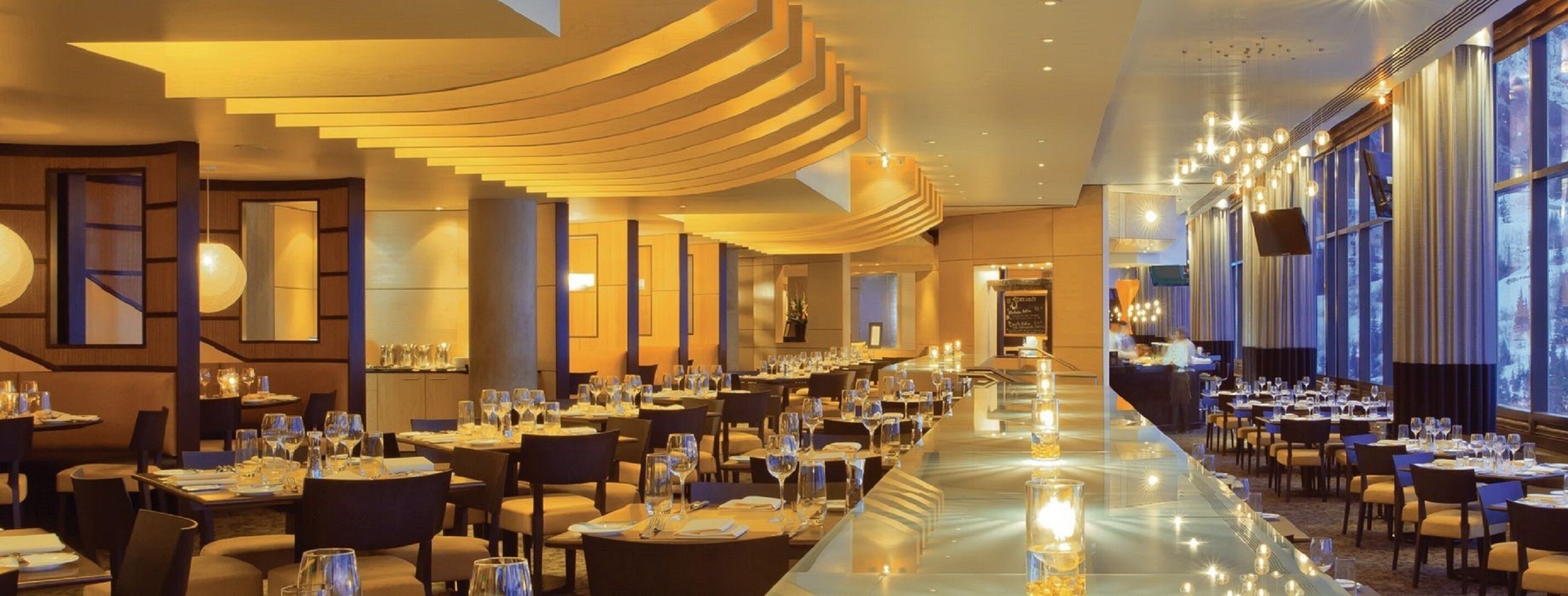 A long dining area with tables set for a meal, a central staircase, and large windows on one side.