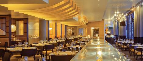 A long dining area with tables set for a meal, a central staircase, and large windows on one side.
