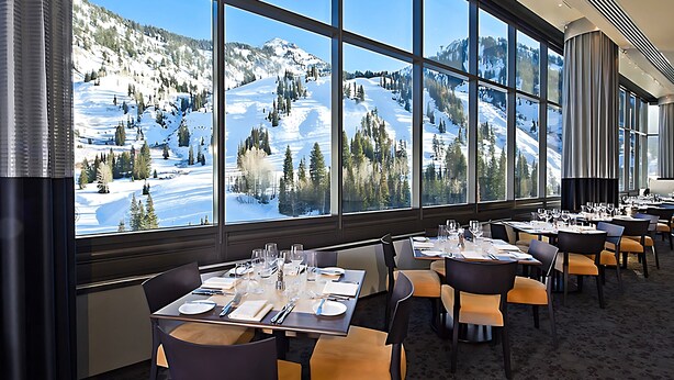 Ein Restaurant mit gedeckten Tischen und großen Fenstern, durch die sich eine verschneite Berglandschaft blickt.