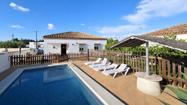 A swimming pool with lounge chairs, a wooden fence, and a building with a tiled roof. - Chalet rustic Roche with pool (CONIL DE LA FRONTERA)