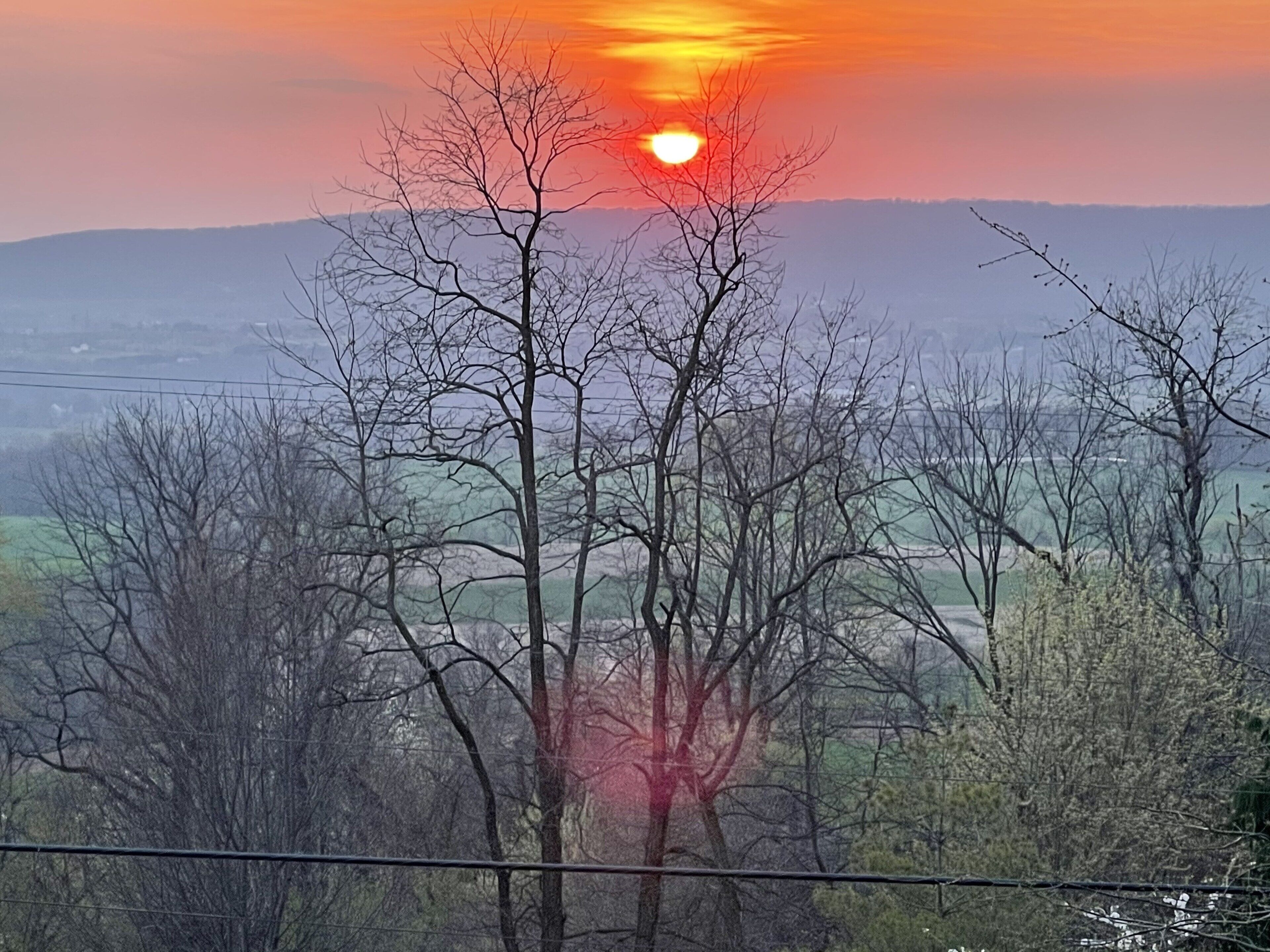 A landscape with a sunset, trees, and a distant view of a town.