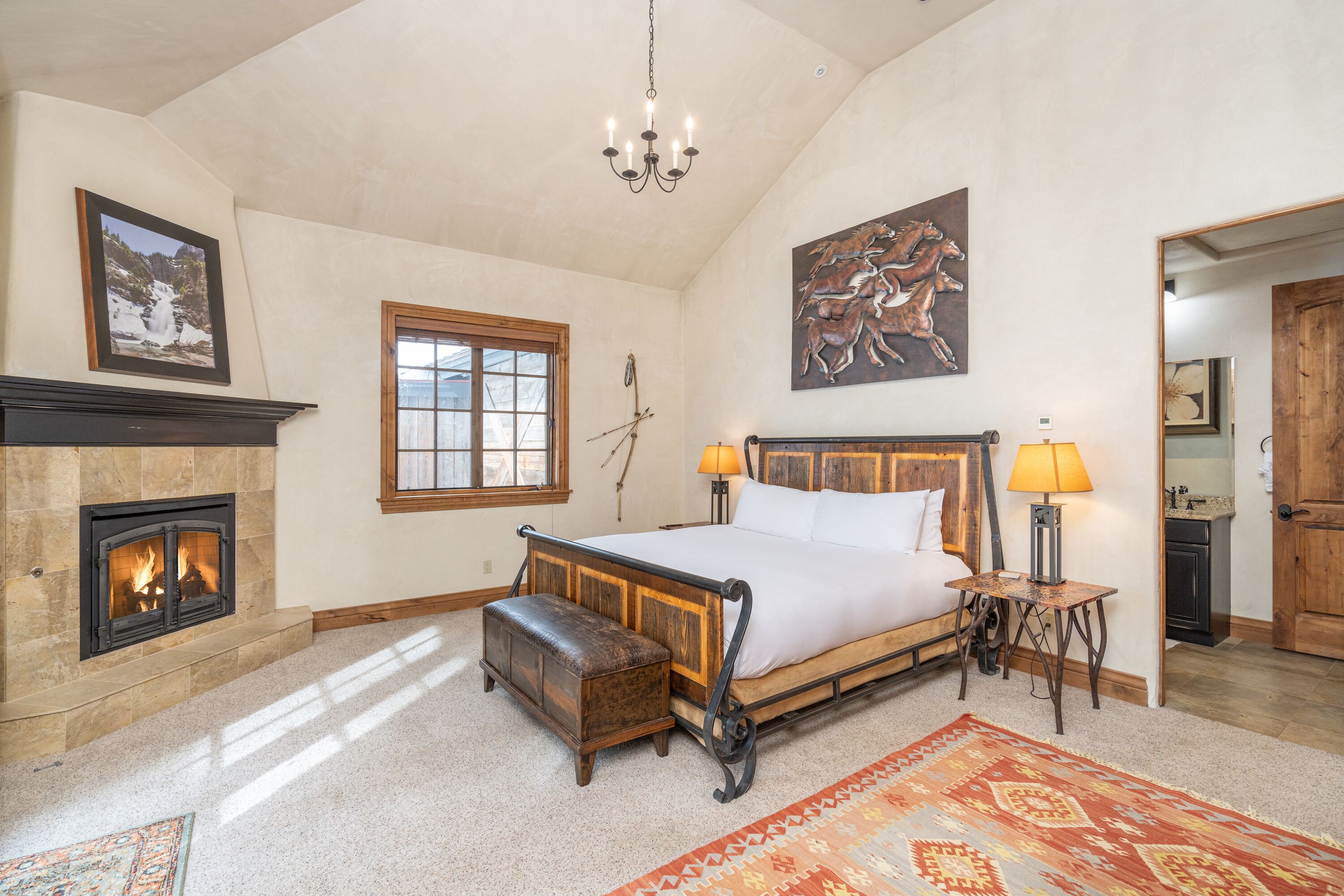A spacious bedroom with a large bed, a fireplace, a chandelier, and a wooden door.