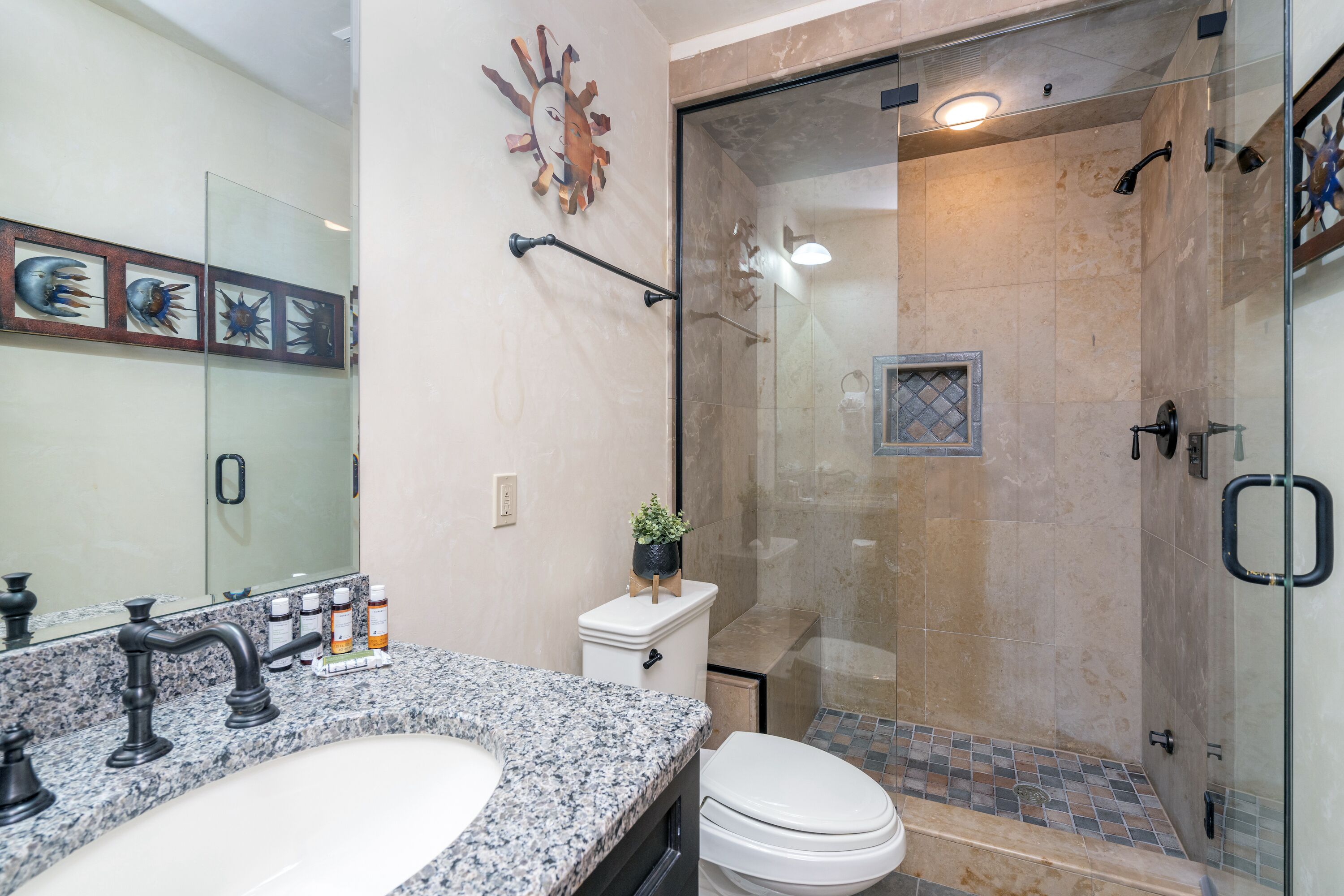 Un baño con encimera de granito, un lavabo blanco, inodoro y zona de ducha con mampara de cristal.