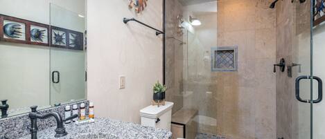 Un baño con encimera de granito, un lavabo blanco, inodoro y zona de ducha con mampara de cristal.