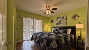 Ein Schlafzimmer mit einem Bett, einem Deckenventilator, einem Nachttisch, einer Lampe und einem Fenster mit Jalousien.