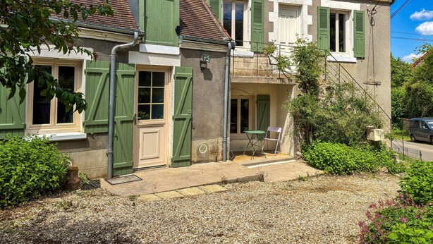 Une maison à deux étages avec une allée en gravier, un petit porche doté d’une chaise et une fenêtre avec des volets.