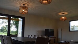 Een moderne eetruimte met een grote tafel en stoelen, twee hangende hanglampen, een televisie en een raam met uitzicht op de omgeving.
