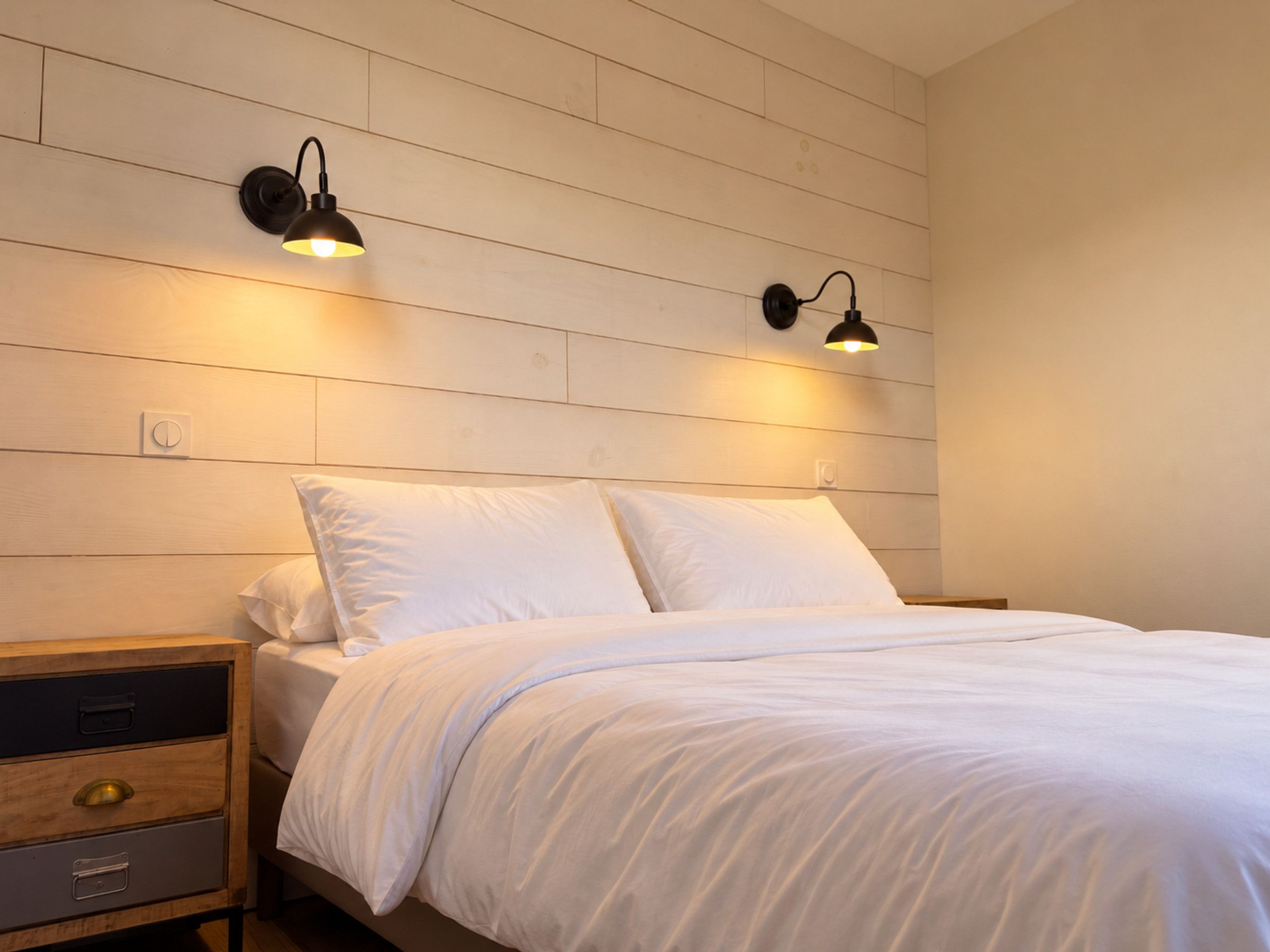 A bedroom with a wooden headboard, two bedside lamps, a wooden nightstand with drawers, and a bed with white bedding.