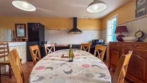 A dining area with a round table, chairs, pendant lights, a kitchen in the background, and a clock on the wall.