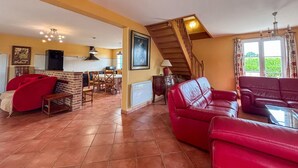 A spacious living area with a red sofa, a red armchair, and a dining table. There is a kitchen with a stove and a refrigerator. A staircase leads to an upper level.