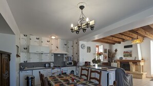 A kitchen with white cabinets, a dining table with a fruit-patterned tablecloth, wooden chairs, a chandelier, and a fireplace. - Charming Stone House with Terrace, Kids’ Playground, and Equestrian Space in Scenic Berry (Pérassay)