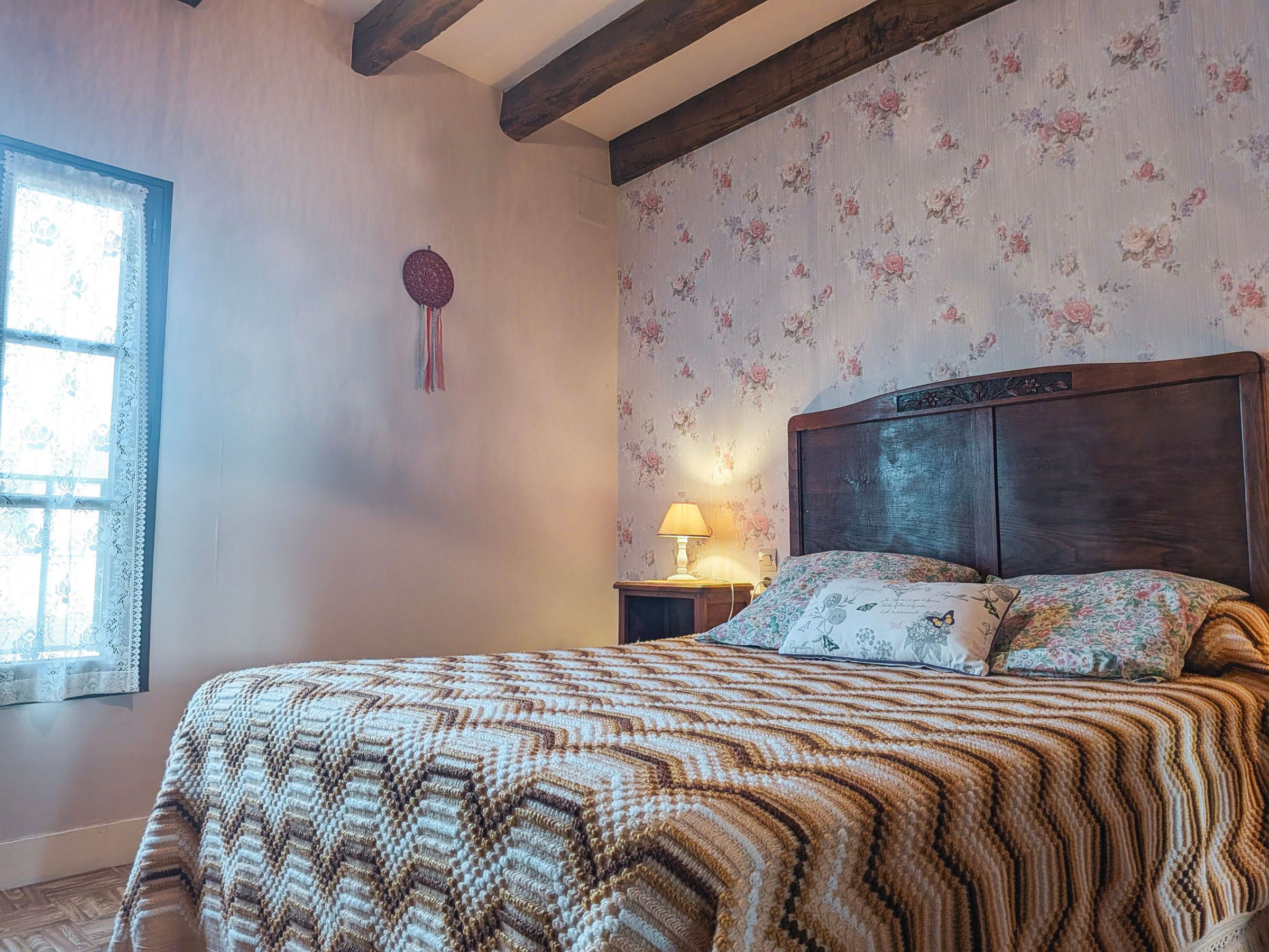 A bedroom with a wooden headboard, floral wallpaper, a patterned bedspread, a bedside lamp, and a window with lace curtains.