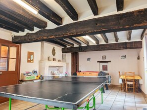 A ping pong table in a room with exposed wooden beams, a fireplace, and a dining area with chairs.