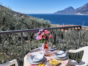 Una mesa dispuesta con platos, una botella y un florero con flores, en un balcón con vistas a un paisaje costero.