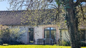 Ein Gebäude mit Ziegeldach, Backsteinwänden und einem großen Fenster. Vorne befindet sich eine Rasenfläche und zur Rechten ein Baum.
