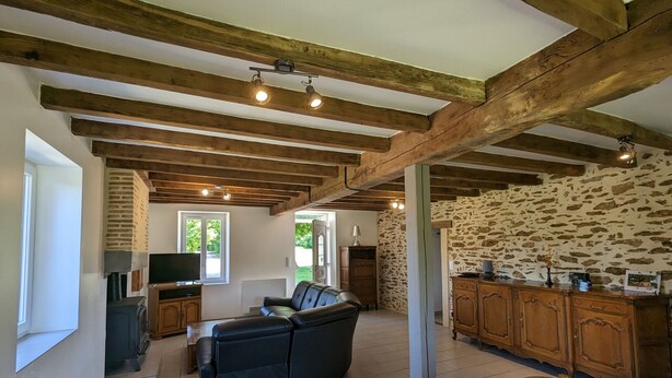 A rustic interior with exposed wooden beams, a stone wall, a black leather sofa, a dining table with chairs, and a kitchen area with wooden cabinets. - Countryside Retreat with Private Garden - Perfect for Family Relaxation! (Pouligny-Notre-Dame)