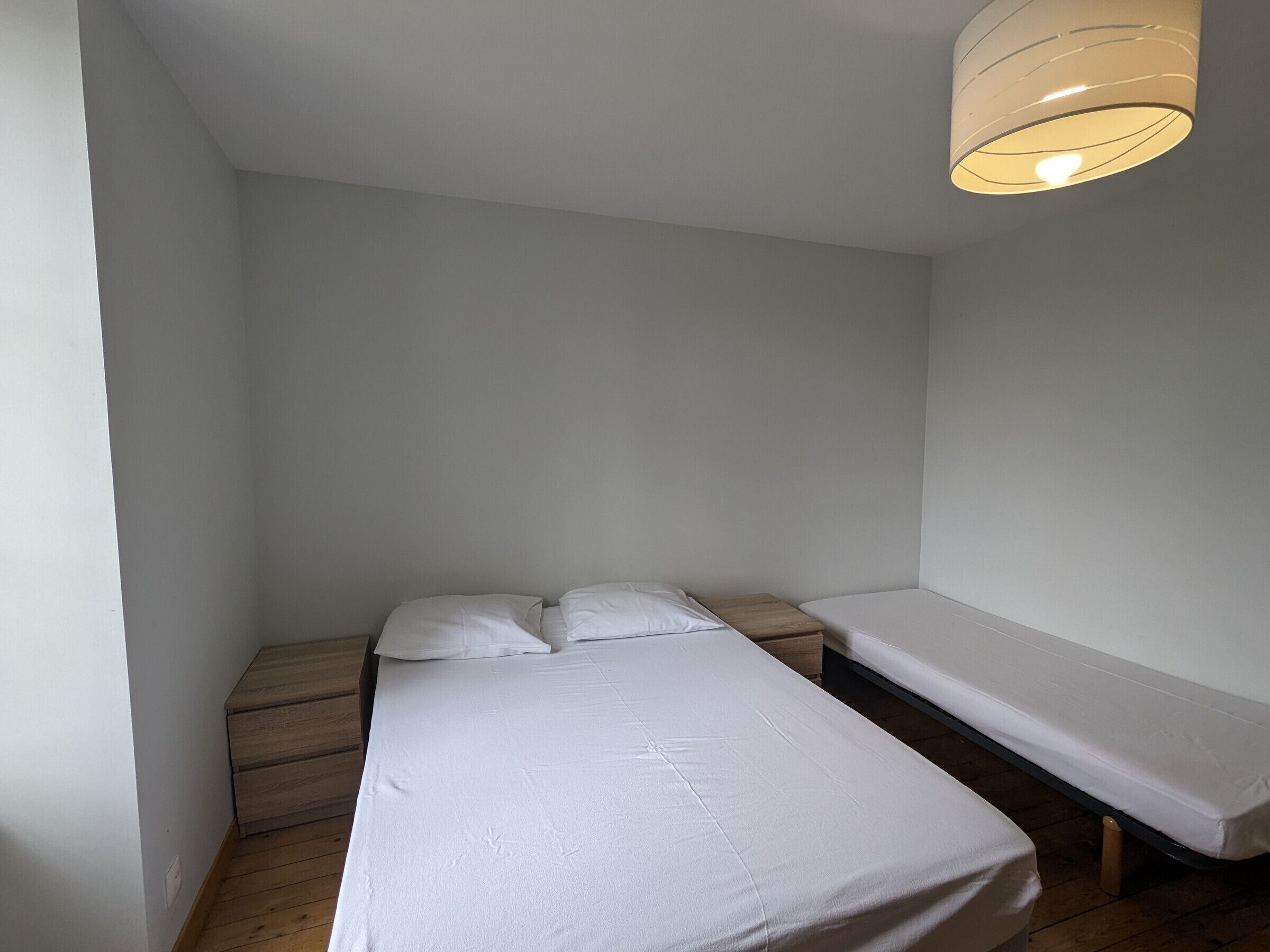 Un petit logement composé d’une seule pièce, comprenant deux lits simples, un plancher en bois et un luminaire au plafond.