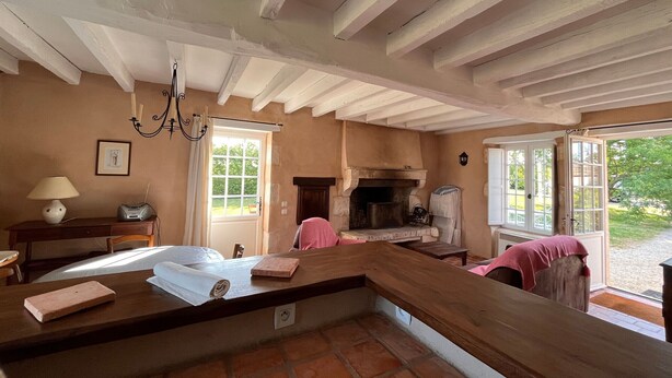 A rustic kitchen with exposed beams, a fireplace, and a view of the garden through large windows. - Peaceful cottage in Berry, family ideal (Sainte-Fauste)