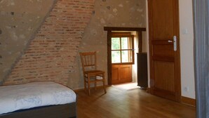 Ein Zimmer mit schrägem Dach, einer Holztür, einem Stuhl und einem Bett mit gemustertem Überwurf.