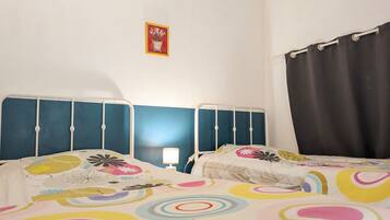 A room with two beds, each with a headboard and a bedside table. There is a wall-mounted picture and a curtain on the right side.