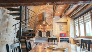 A rustic interior with exposed brick walls, a wooden staircase, a central fireplace, and a dining area with a long table. - Cozy Renovated Forge with Sauna, Shared Pool & Nearby Attractions (Le Menoux)