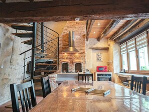 Ein rustikales Interieur mit freiliegenden Backsteinwänden, einer Holztreppe, einem zentralen Kamin und einem Essbereich mit einem langen Tisch.
