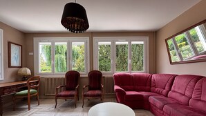 Un salon avec un canapé rouge, deux fauteuils, une table basse blanche et une lampe suspendue. De grandes fenêtres offrent une vue sur la verdure environnante.