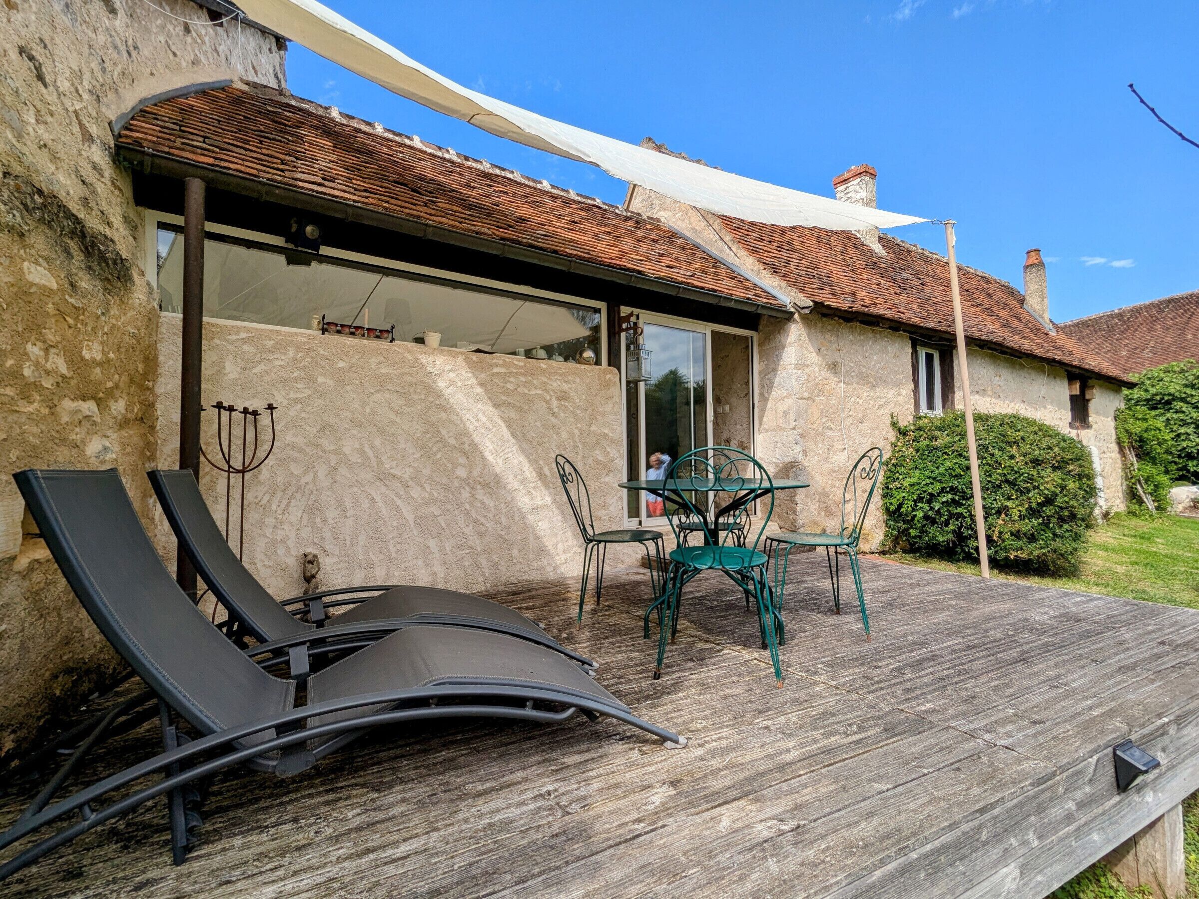 Un bâtiment rustique en pierre, doté d’une terrasse en bois, d’une porte vitrée donnant sur un patio, et d’une table avec des chaises.