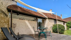 A rustic stone building with a wooden deck, a glass door leading to a patio, and a table with chairs. - Charming Nature Retreat with King Bed, Private Terrace, and Shared Pool in the Brenne Park (Chalais)
