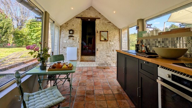 A kitchen with stone walls, a dining area with a table and chairs, and a view of the garden. - Charming Nature Retreat with King Bed, Private Terrace, and Shared Pool in the Brenne Park (Chalais)