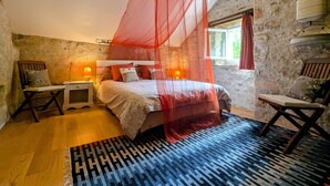 A bedroom with a stone wall, a wooden bed with a red canopy, a nightstand, a chair, and a window with curtains. - Charming Nature Retreat with King Bed, Private Terrace, and Shared Pool in the Brenne Park (Chalais)