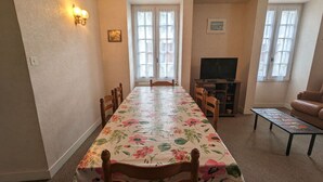 Et spiseområde med et langt bord dækket af en blomstret dug, omgivet af træstole. Et fjernsyn er monteret på et stativ til højre, og en brun lædersofa står placeret i hjørnet.