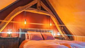 Un dormitorio acogedor en el ático con techo inclinado, vigas de madera a la vista, una cama grande con almohadas y dos lámparas de noche.