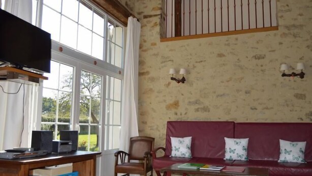 Una habitación con un escritorio de madera, televisión, un sofá rojo con cojines estampados, una silla y un ventanal con cortinas.